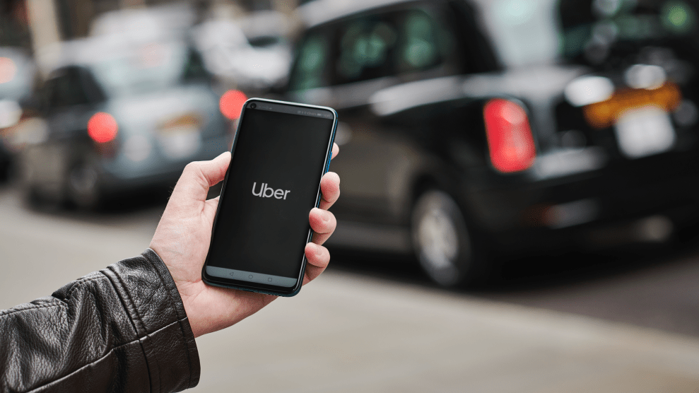 A hand in a black leather jacket uses the Uber app on a smartphone with a black London taxi in the background.