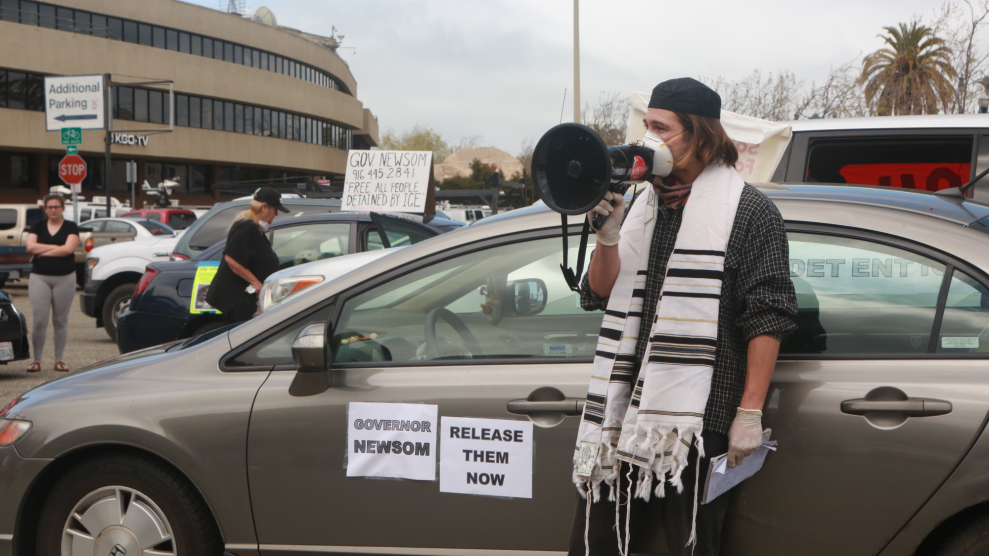 Wearing mask and gloves, plus a kippah and other Jewish garments, person standing outside of a car speaks through a megaphone.