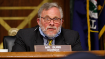 Sen. Richard Burr, with a gray beard and glasses, speaking behind a nameplate in a Senate chamber, with a mask pulled down around his chin.