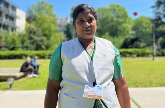 A photo of a woman with brown skin and dark brown hair wearing a green short and gray vest outside.