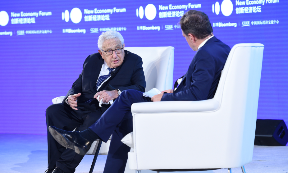 Henry Kissinger seated onstage in an interview wearing a dark suit on a white chair.