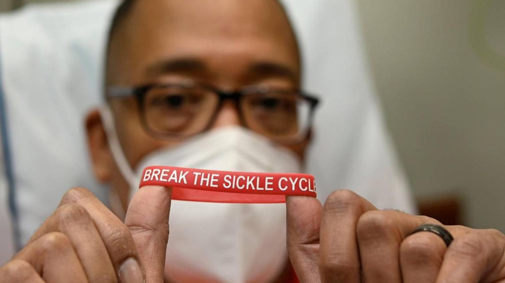 A Black man wearing glasses and a white N95 mask sitting in a hospital bed holding a red bracelet that says "Break the Sickle Cycle"