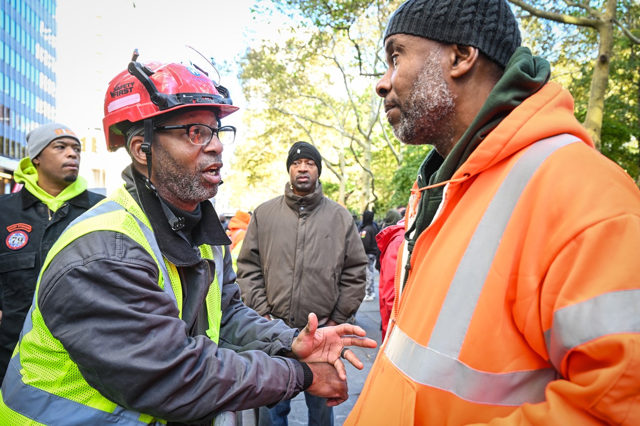 A man in a red hart had speaks to another man in an orange safety vest.