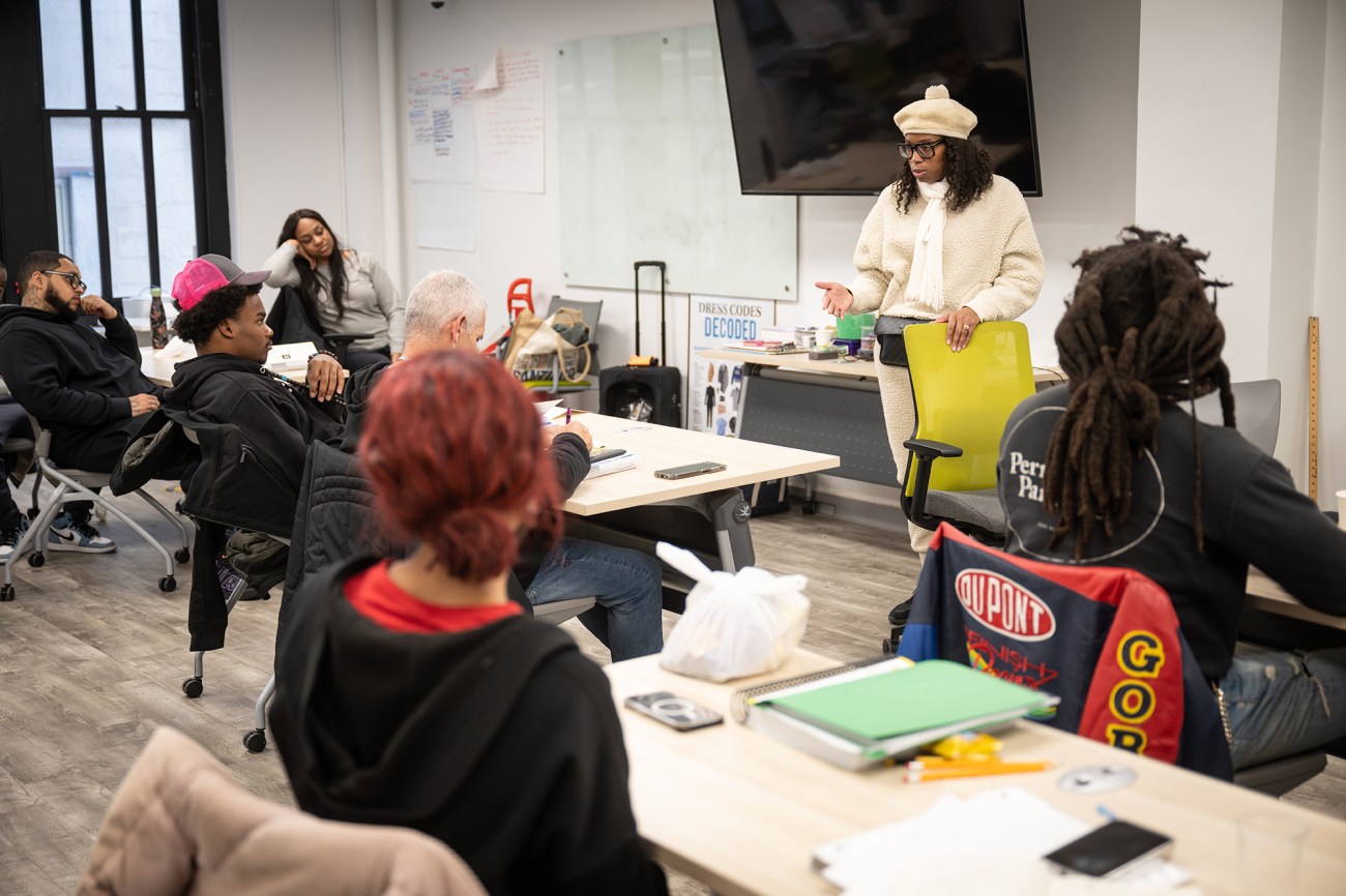 Woman in a white hat and sweater stands, talking in front of a classroom.