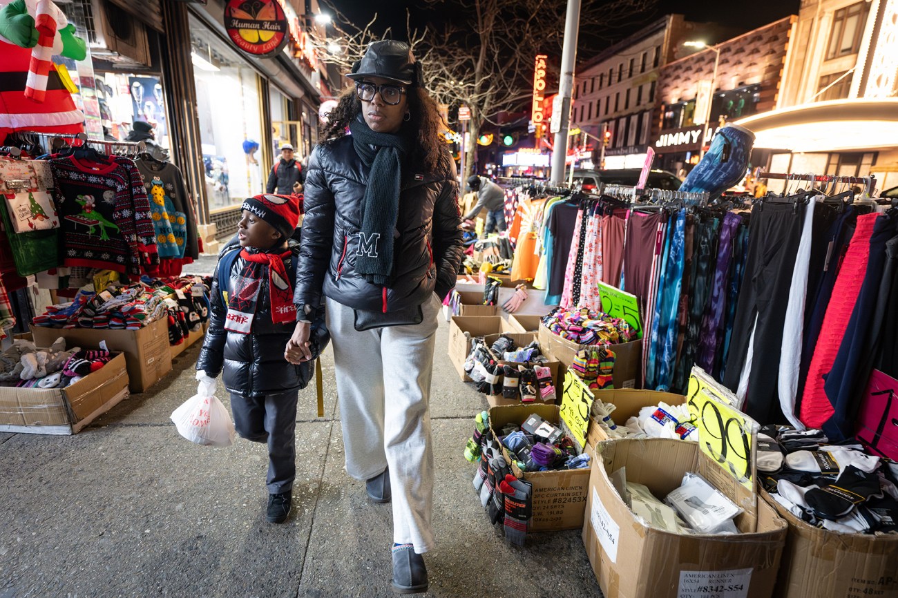 A woman wearing a black jacket walks down the street holding the hand of her child.