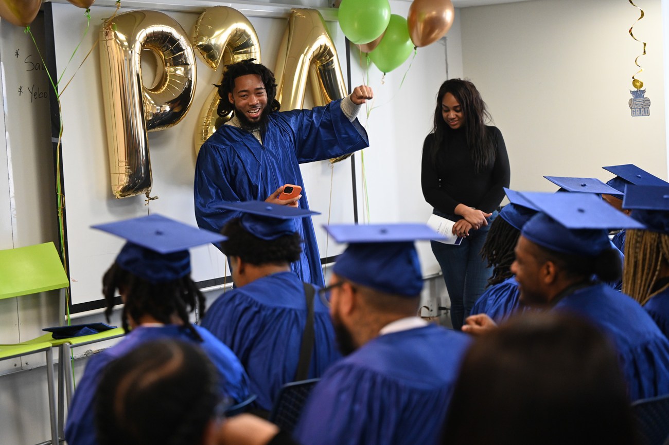 A smiling man wearing a blue graduation gown raises his fist in front of other graduates.