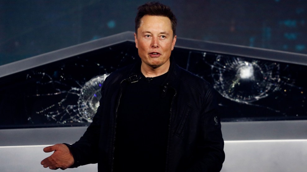 A white man in a black outfit stands in front of a damaged vehicle.