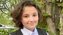 Photo of a white trans teen with brown hair wearing a white button down and black vest.