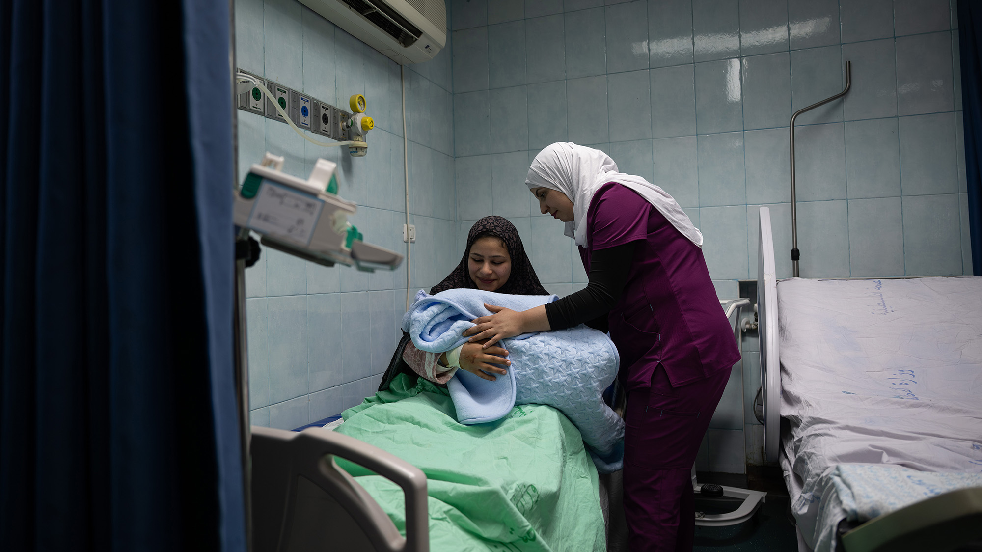 Woman sitting in hospital bed holds her newborn. A midwife stands over her.