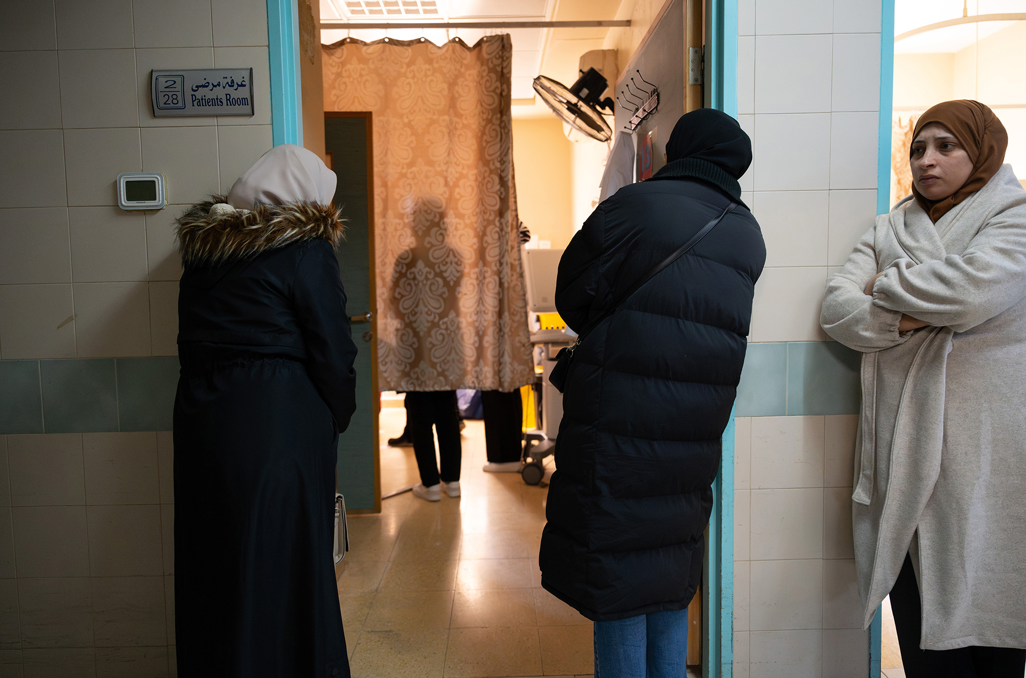 Three women stand around the door to a hospital room door, two of them looking into the room.