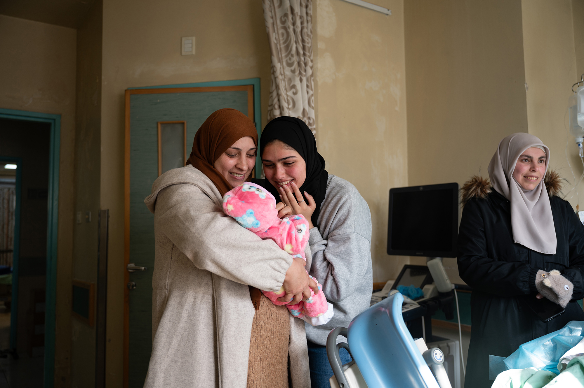 Two women look at a baby wrapped in a blanket. A third woman is smiling, looking out of the right of the frame.