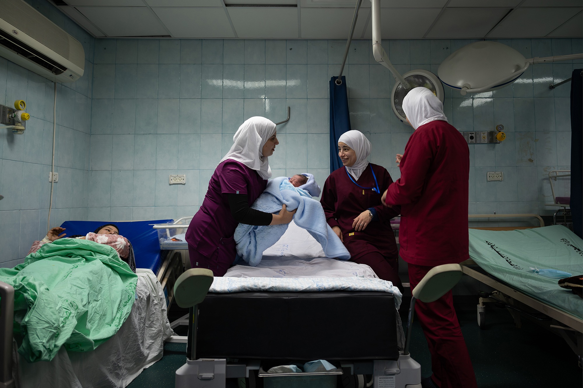 Three women stand in a hospital operating room, one holding a newborn baby. A woman lays on a hospital bed, watching.