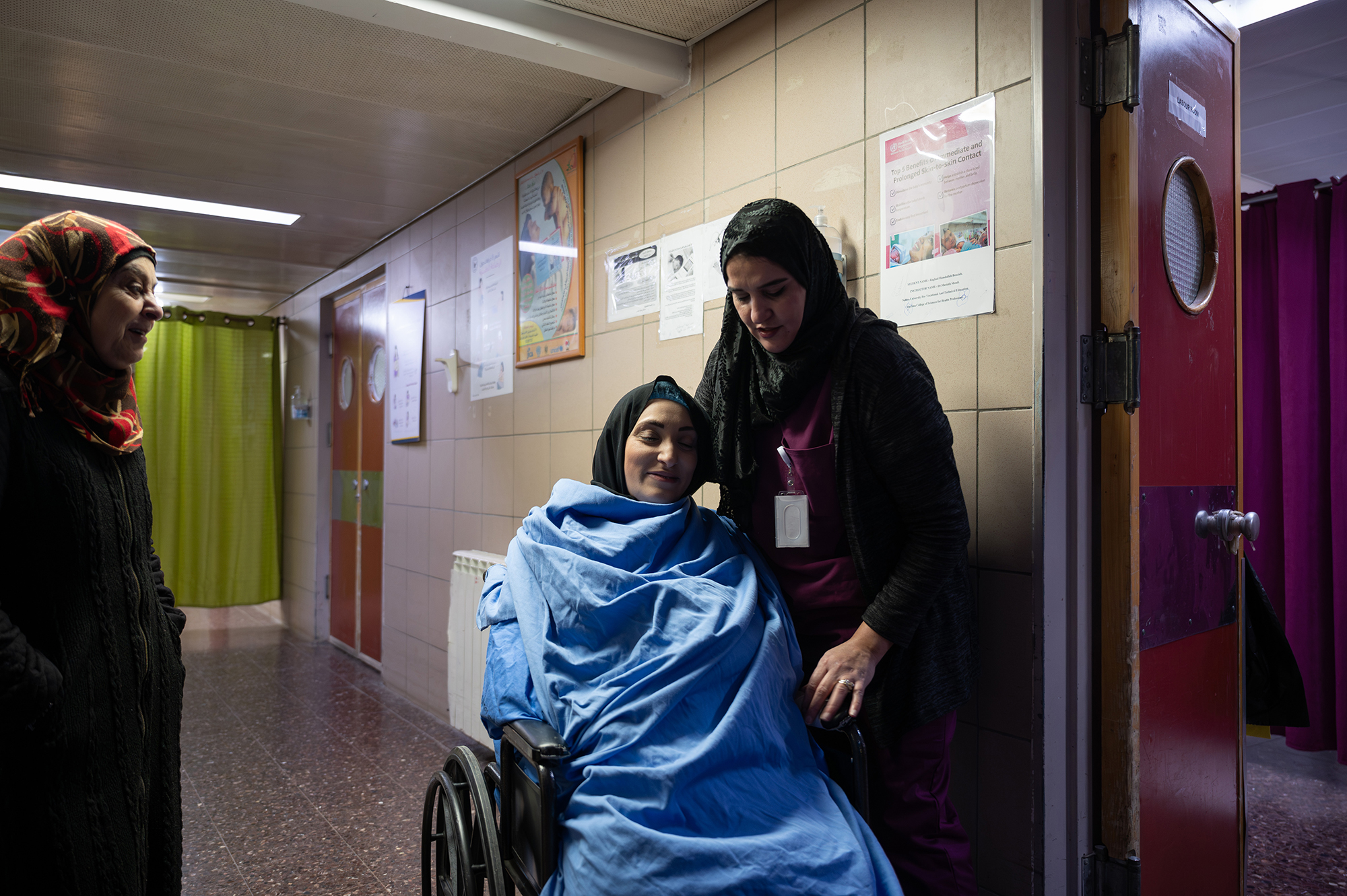 A woman with her eyes closed, sitting in a wheelchair in a hospital. Another woman is standing next to her. A third woman looks on from the left of the frame.