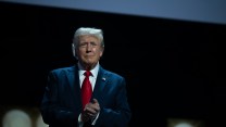 Donald Trump in a suit with a red tie and bandage over his ear standing on a stage