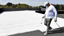 Man standing on roof, spraying it white.