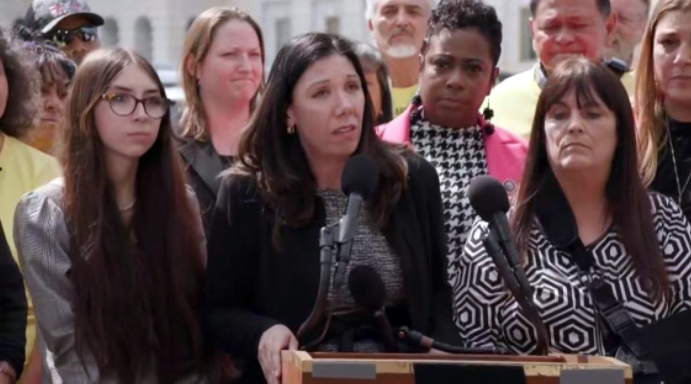 Woman speaking at a lecturn, surrounded by other people.