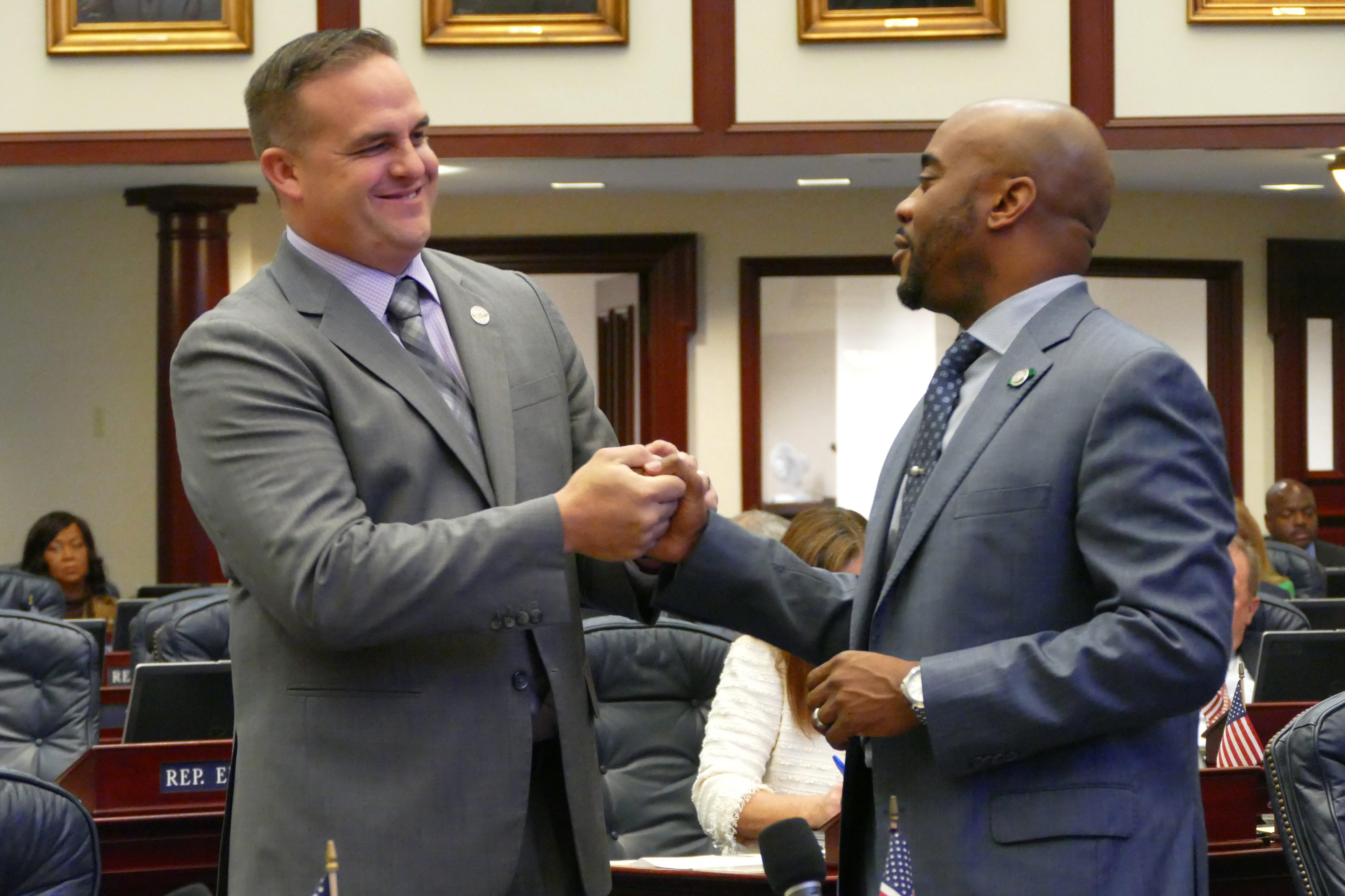 Two men in grey suits smile and shake hands.