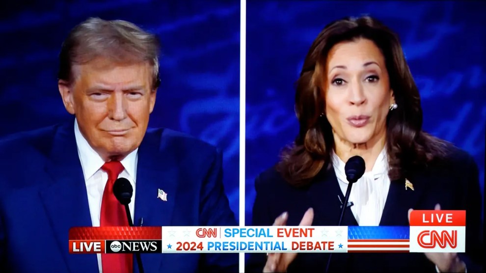 a split-screen view of a live televised 2024 presidential debate between Donald Trump and Kamala Harris. Trump is on the left side of the screen wearing a dark suit with a red tie, and Harris is on the right side, also in a dark suit, speaking into a microphone.