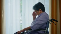 An aging person with light brown skin and dark gray hair in a wheelchair before the window. They have one hand over their face and are looking down. They are wearing a blue shirt.