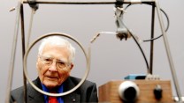 James Lovelock's face is framed by a machine that has wires and pipes swirling around.