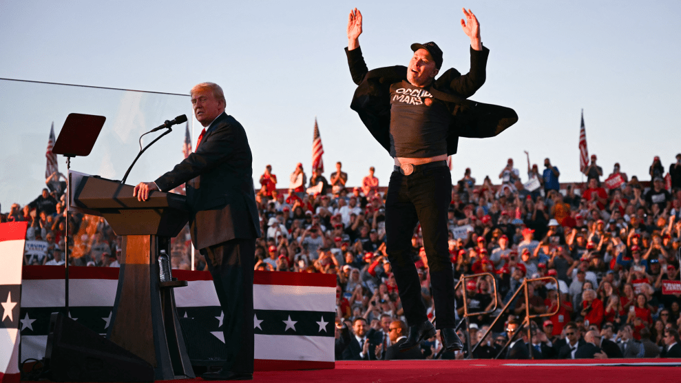 Elon Musk, dressed in all black, on stage behind Donald Trump. As Musk descends from his leap, his arms are raised in mid air, his jacket billowing in the wind.