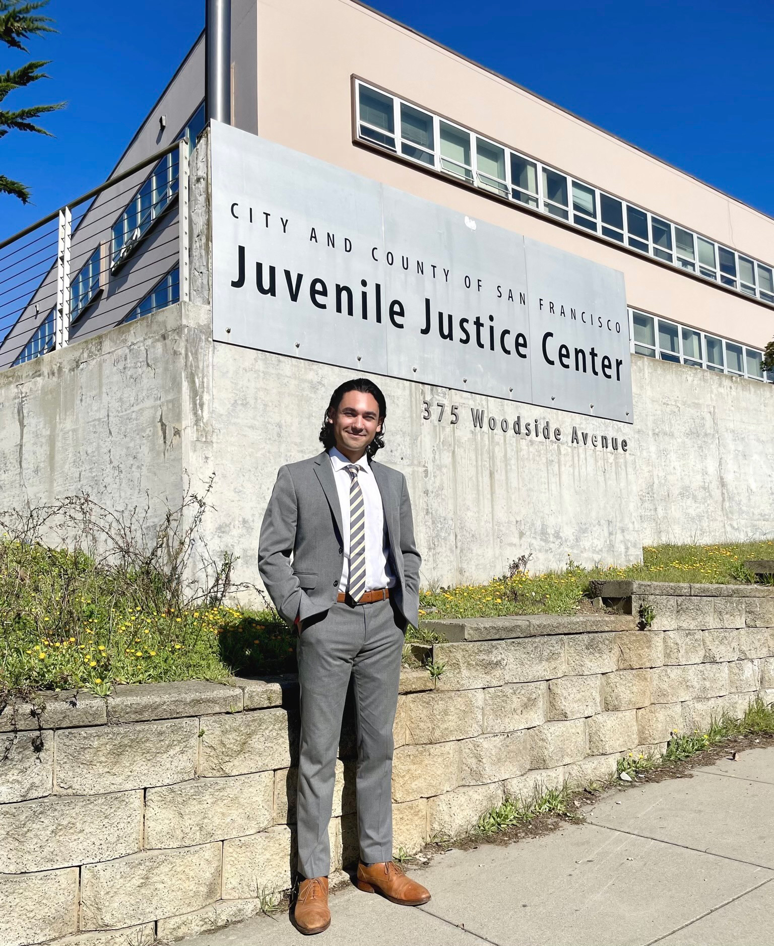 Ryan Khojasteh, dressed in gray suit and striped tie, stands in front of the San Francisco Juvenile Justice Center on a bright, sunny day.