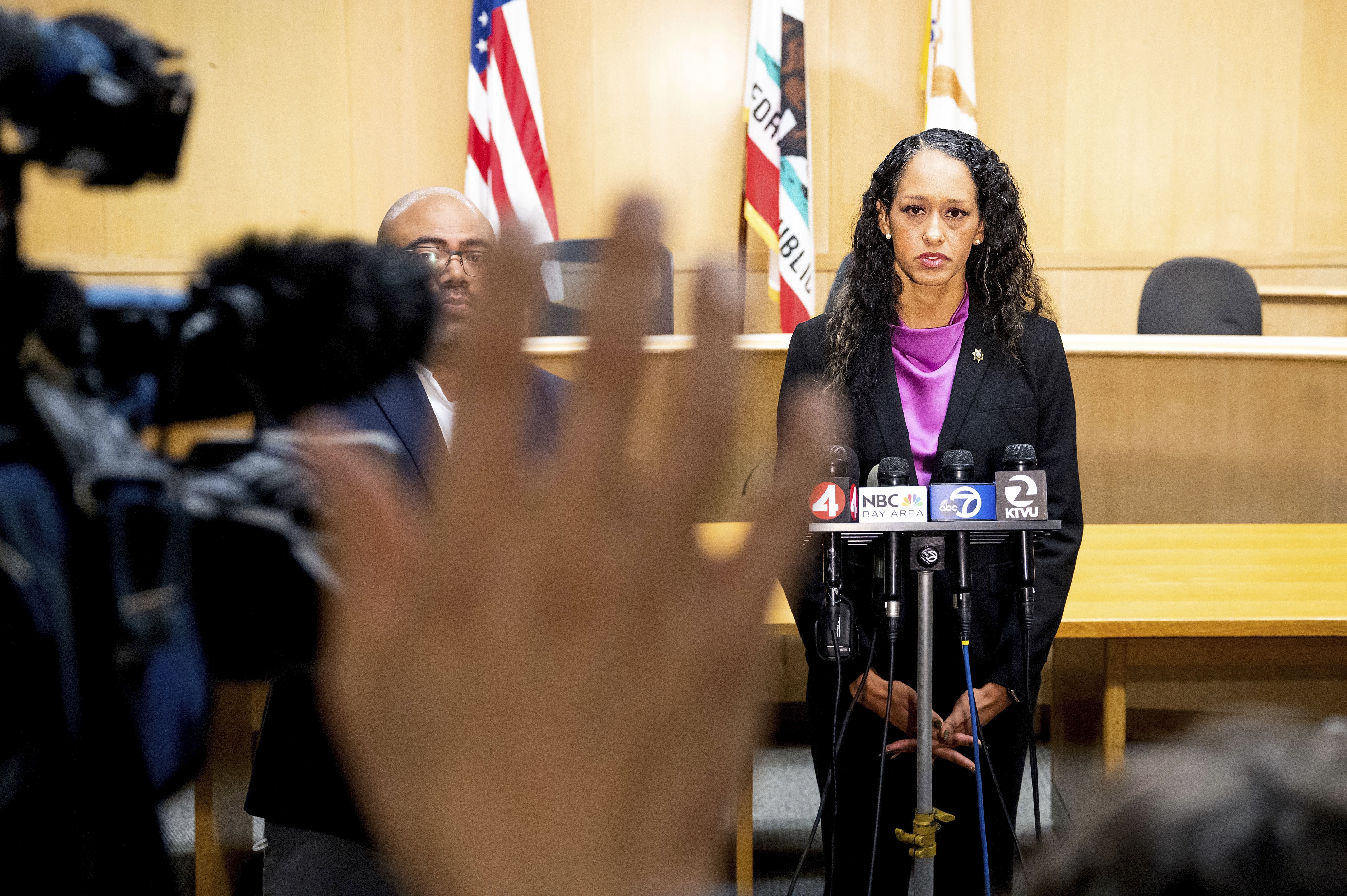 Brooke Jenkins stands in front of a bank of microphones. On the left, we see an out-of-focus raised hand from a member of the media.