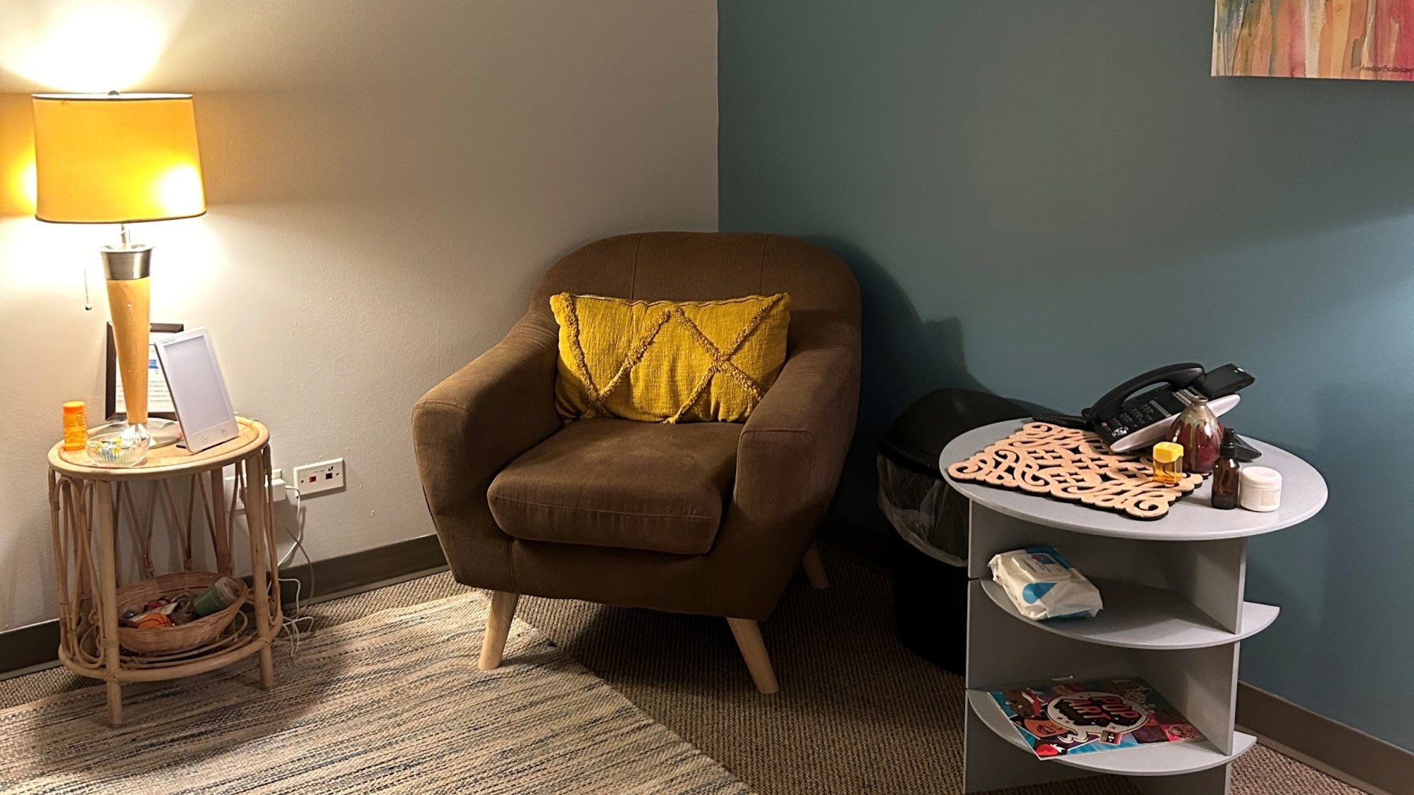 A carpeted room with an upholstered chair and two end tables. A lamp sits on one table and a phone on the other.