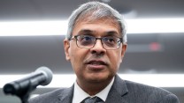 A close-up of Jay Bhattacharya wearing glasses and a grey suite and tie, speaking into a microphone.