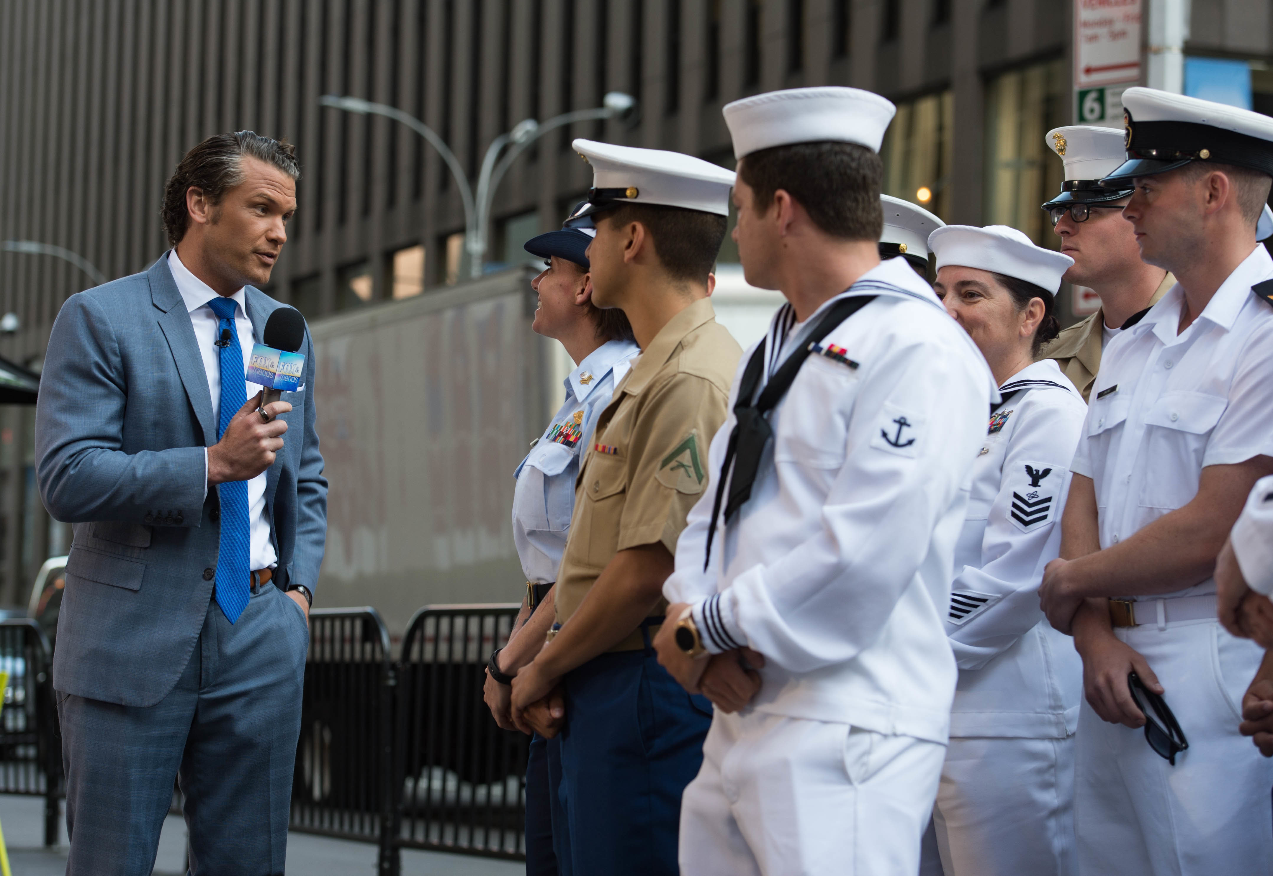 Pete Hegseth holds a microphone, facing a group of uniformed service members.