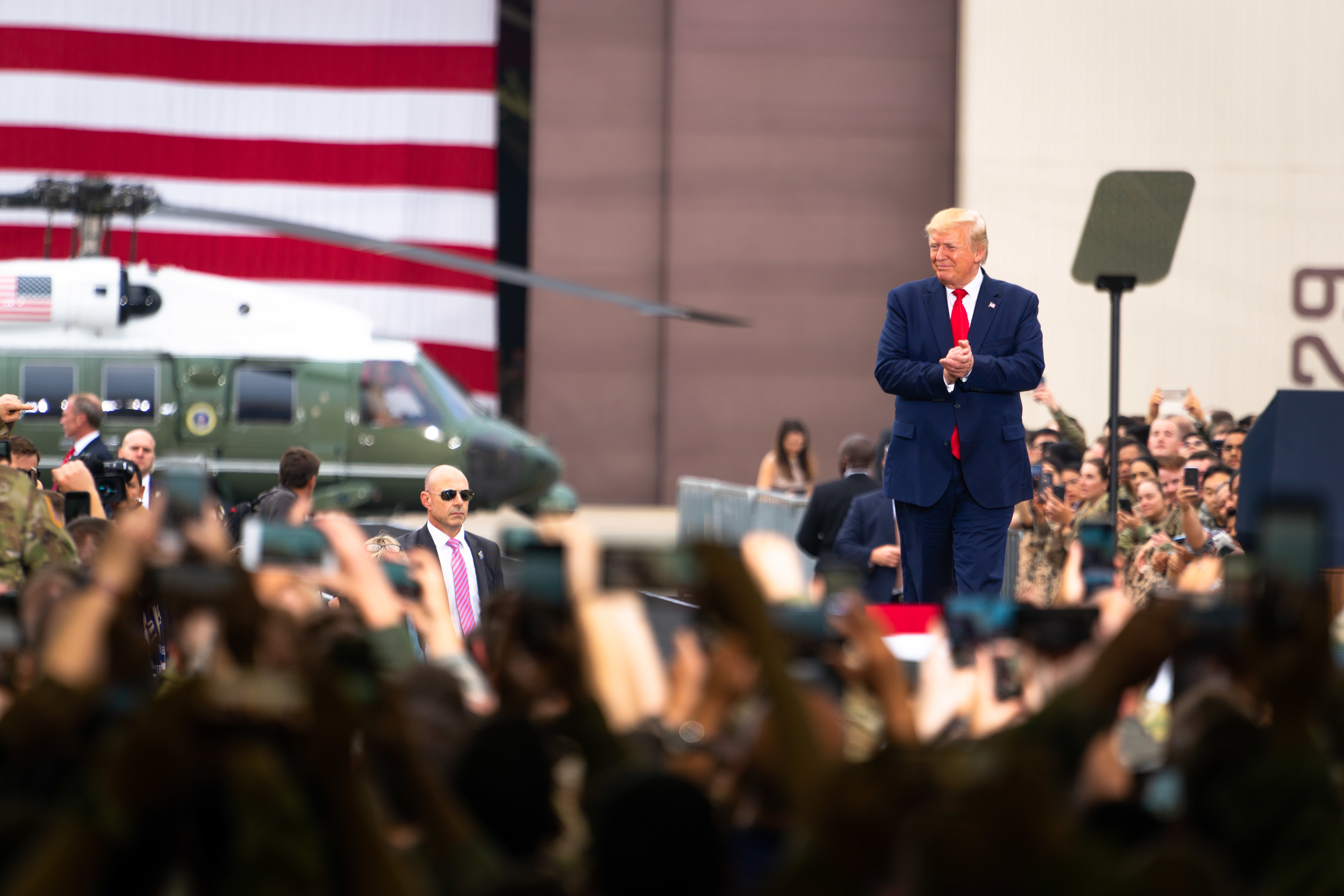 Service members hold up cellphones as Donald Trump walks down an aisle between them. A military helicopter stands in the background.