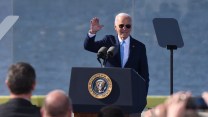 A white man in a suit with a blue tie raises his hand while addressing a crowd.