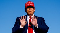 President Donald Trump raises his hands to the camera; he is wearing a Make America Great Again hat and is set against a blue sky.