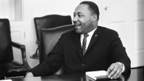 A Black man smiles. He wears a suit. The photo is in black and white.