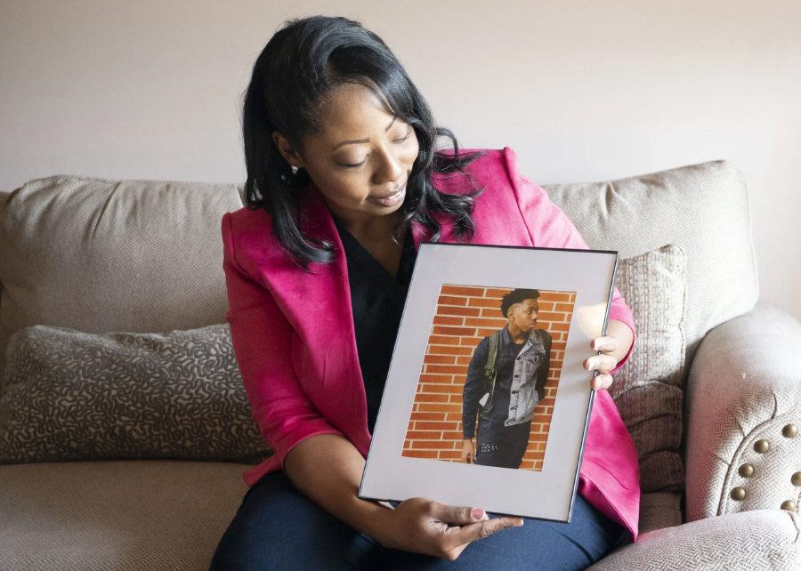 Candace Leslie, a black woman, sits on a couch in her living room, looking at the framed photo of her late son, Cameron Brown, that she holds in here hands.