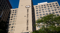 Pictured is a large-brown-bricked building in the Modernist architectural style of the mid 20th century. It's a rectilinear building with repetitive window patterns, and it appears to tower up the sky. There is a large sign that reads 'VA MEDICAL CENTER,' which is positioned vertically down the side of the building. There are dark green deciduous trees in front of the building, concealing some of the lower windows.