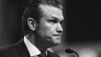 Black and white zoomed in photo of Pete Hegseth; his lips are pursed as he sits in front of a microphone answering questions at a hearing.