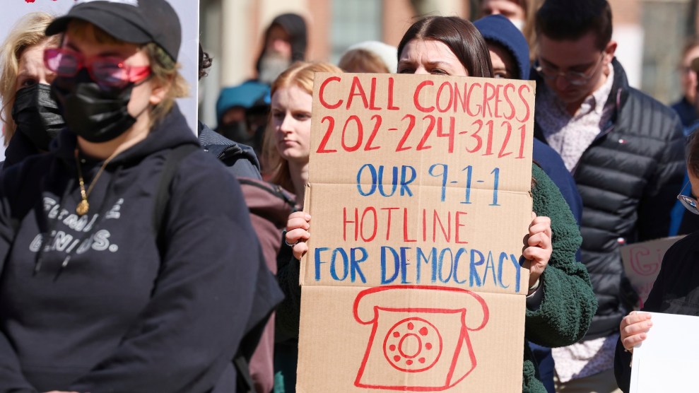 A white woman with brown hair holds up a sign that says "Call Congress 202-224-3121 Our 9-1-1 hotline for Democracy."