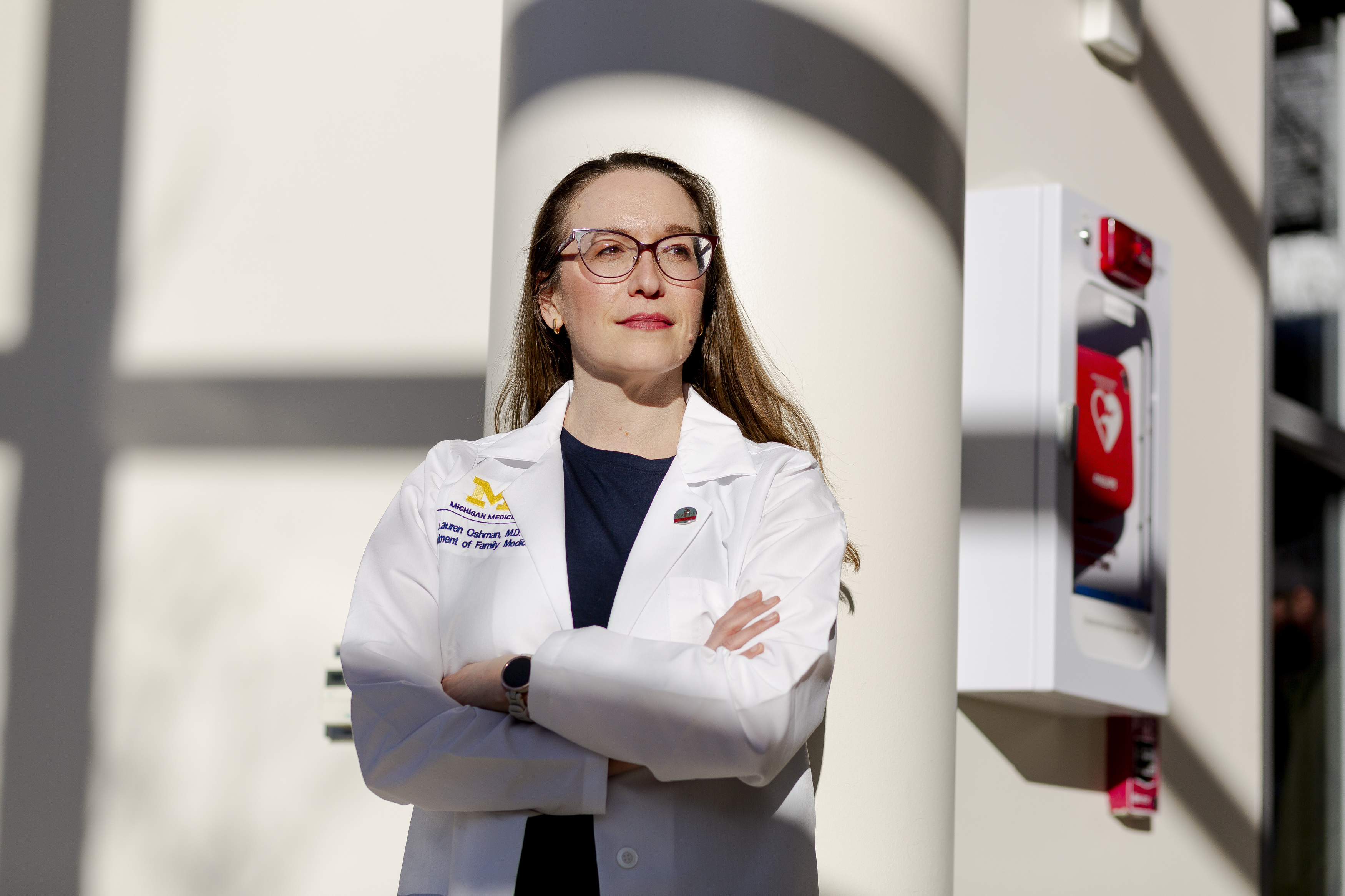 A white female doctor poses for a portrait inside a hospital. She is wearing glasses, a black blouse and a white coat with a label that reads “Michigan medicine, Lauren Oshman, M.D., Department of Family Medicine.”