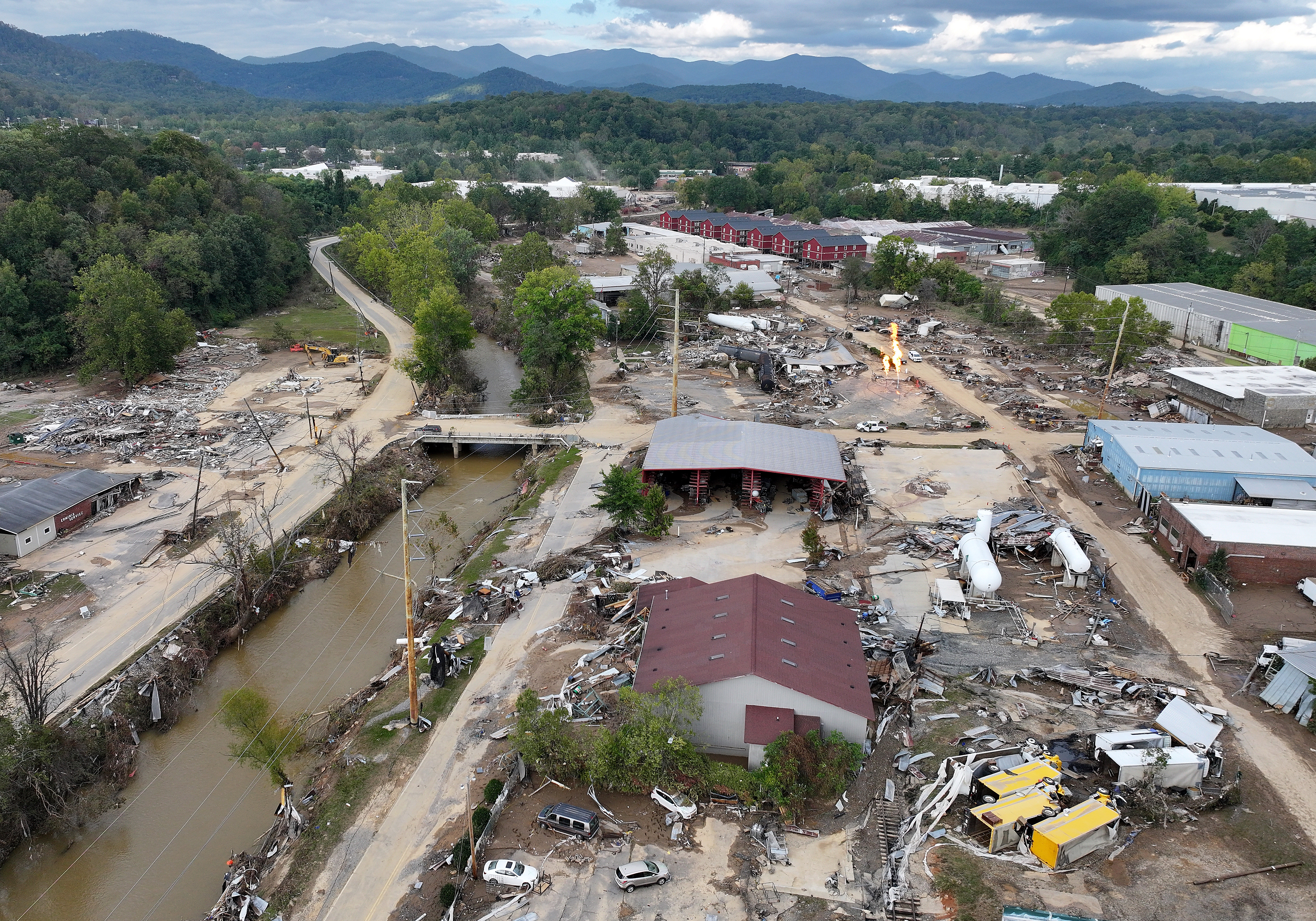 Shown from the air, large piles of construction debris litter a developed riverbank. A few buildings stand among rubble-filled lots where other buildings were destroyed.