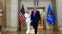 Pam Bondi, dressed in an all-white suit, and Donald Trump, dressed in a navy suit and red tie, walk into a large room with smooth stone walls.