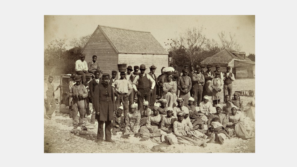 A photo of scores of men and women, mostly black, in mid-19th century clothing and hats pose, looking directly into the camera. Women and children sit together on the ground. Men and a few women stand behind them. In the background are wood structures set against trees. A white Union soldier in full uniform and beard stands in the foreground.