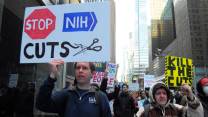 Protesters hold up signs, including one that reads, Stop NIH Cuts, with a stop sign and a pair of scissors