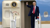 President Donald Trump stands in the doorway of Air Force One airplane, wearing a suit and red tie, holding up one fist.