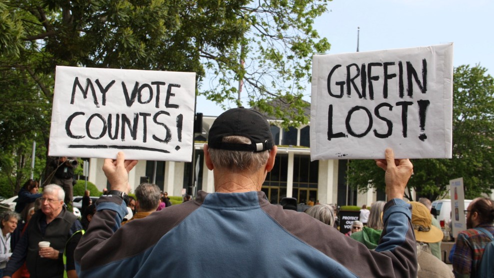 A man at a protest stands with his back to the camera and holds two white signs with blank inking, reading: My vote counts! and Griffin Lost!