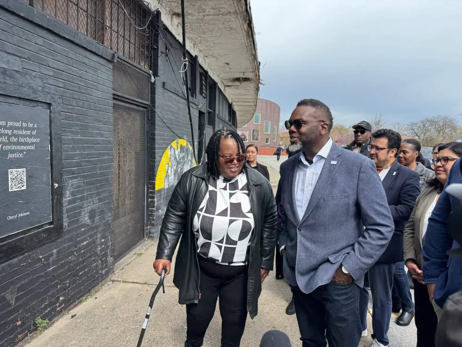 A Black woman with a cane walks with a group of people through a neighborhood street.