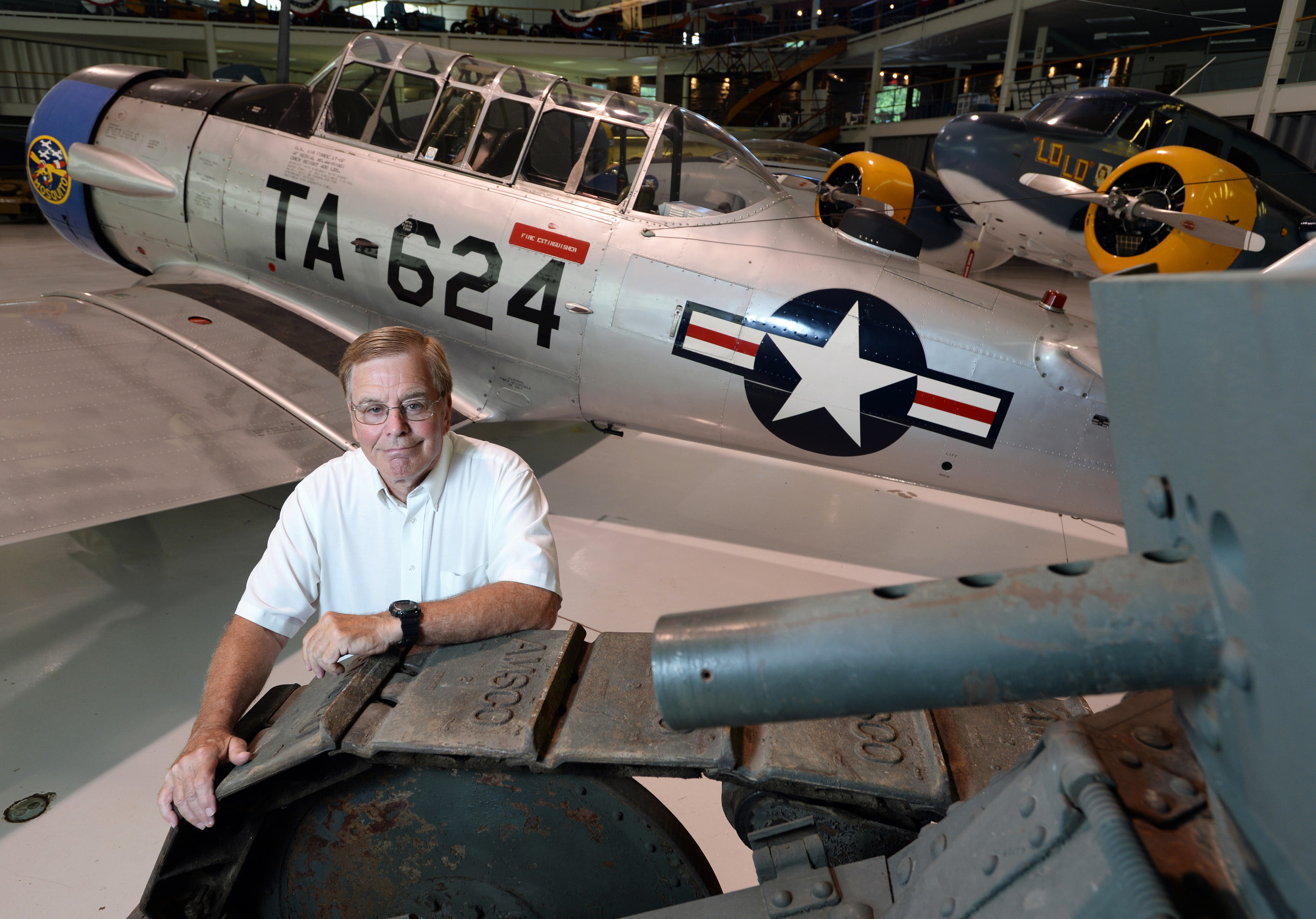 Man standing, leaning against the tracks of an old tank with an old fighter plane behind him.