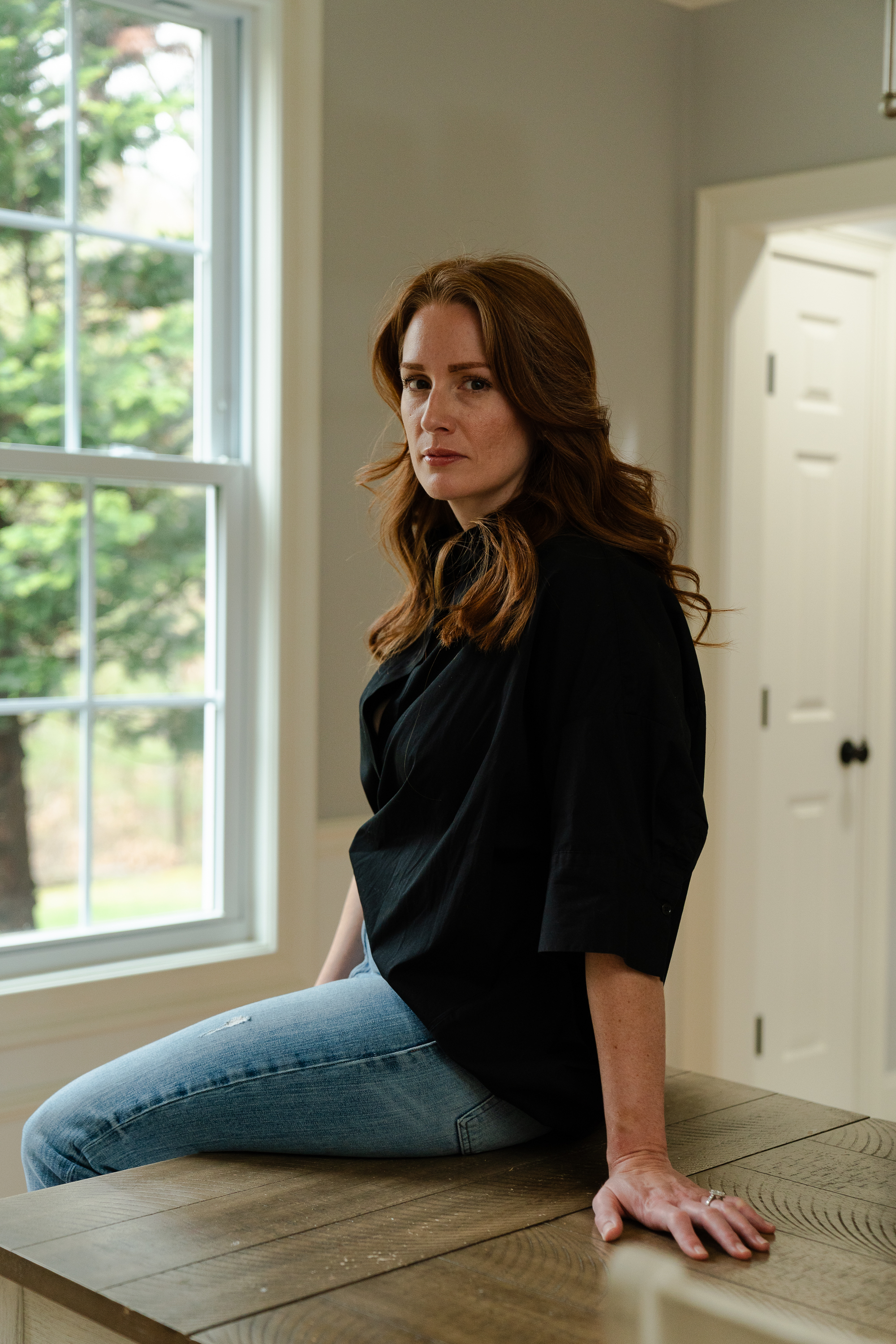 Portrait of red-haired woman sitting on a table near a window.