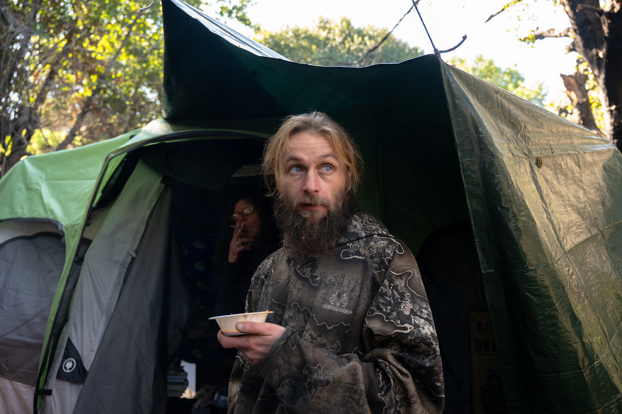 Bearded man holding a bowl of food, standing in front of a tent.