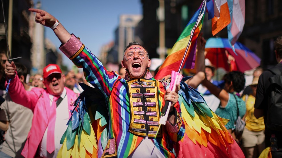 A smiling man walks in front of other celebrants, wearing a long tailed rainbow marching uniform with bejeweled bedazzling.